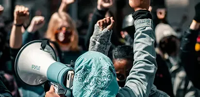 A person in a hijab holding a megaphone in front of a crowd of people with their arms in the air.