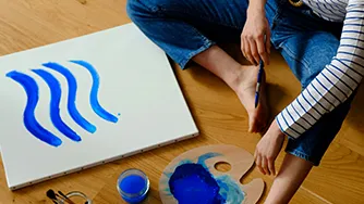 A person sitting on the floor, with a paintbrush in hand, painting a sheet of paper.